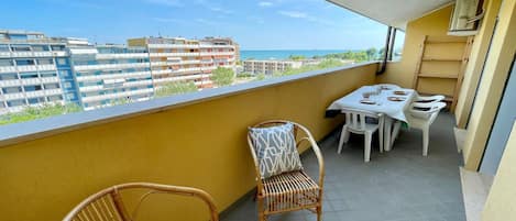 A balcony with a table and chairs, a wicker chair, and a view of buildings and the sea.