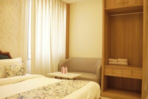 A hotel room with a bed, a chair, a small table with water bottles, and a wooden wardrobe. - The Landmark Maan Palace Hotel (Jaipur)