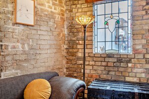 A room with a brick wall, a sofa with cushions, a floor lamp, and a window with a stained glass design. - Host & Stay | The 1890 Petersfield Retreat (Cambridge)