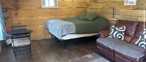 A cozy cabin-style room with a bed, a sofa, a chair, a TV, and a window.