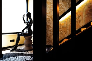 A sculpture of a human figure seated on a pedestal, positioned near a window with a patterned wall in the background. - Lagos Moments Lodge (Lagos)