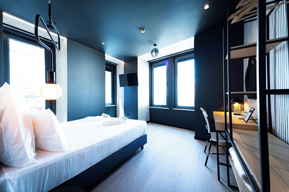 Habitación de hotel moderna con una cama grande, un escritorio y una silla. Hay ventanas con persianas, una lámpara de techo y vistas al exterior.
