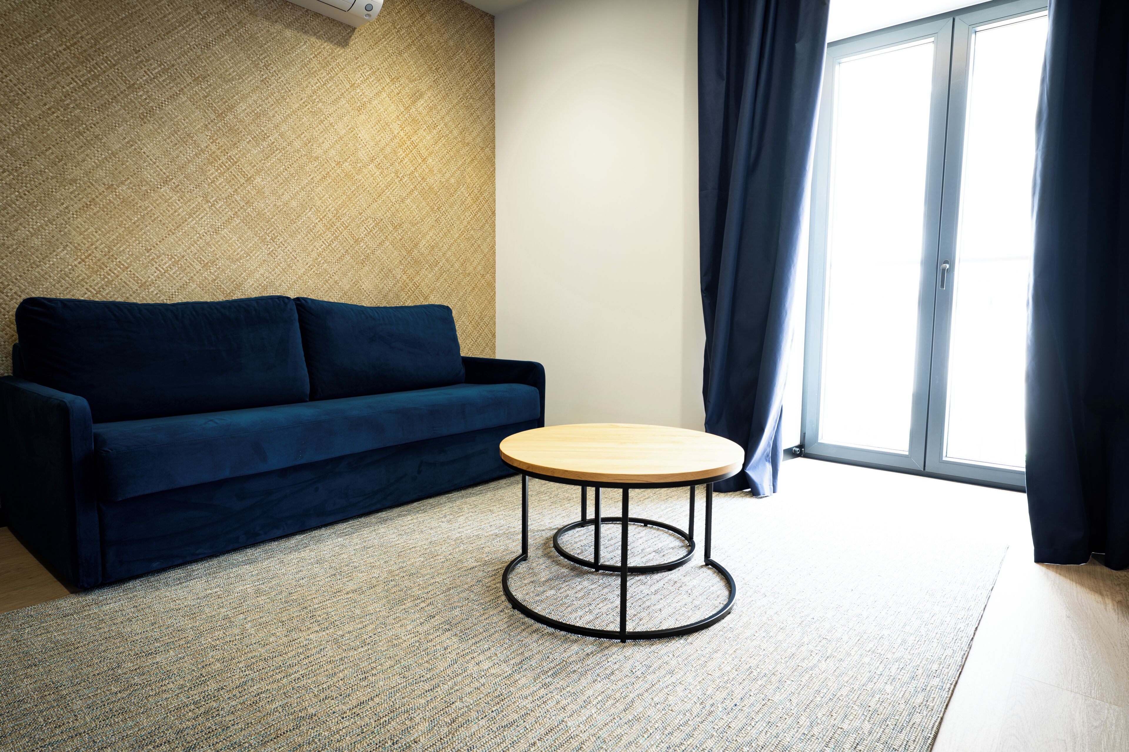 Une pièce avec un canapé bleu foncé, une table ronde en bois avec un pied en métal, un tapis tissé et une grande fenêtre avec des rideaux.