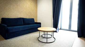 Et værelse med en mørkeblå sofa, et rundt træbord med metalstel, et vævet tæppe og et stort vindue med gardiner.