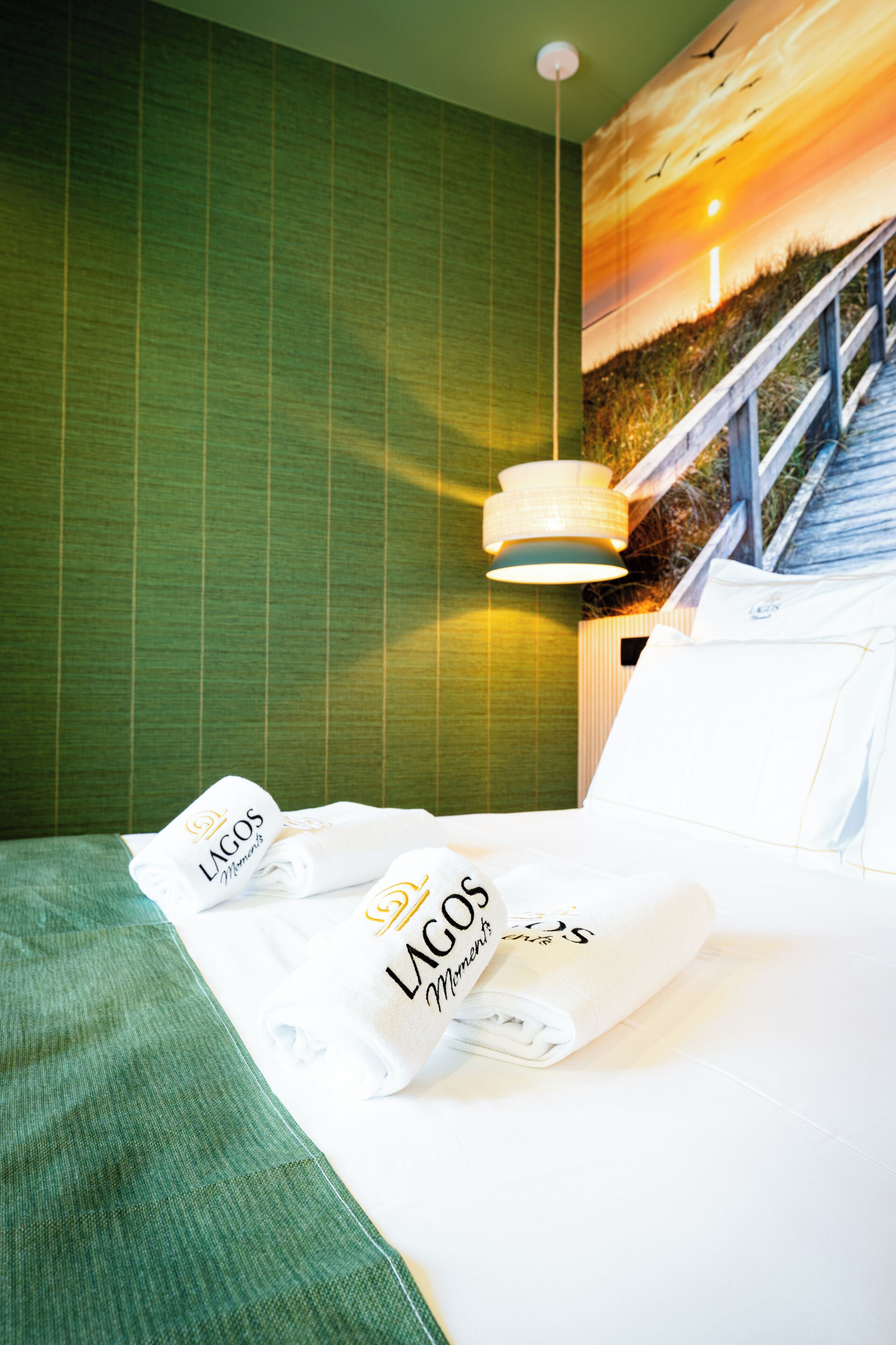 A hotel room with a bed, a green wall, a painting of a sunset, and a hanging light fixture.