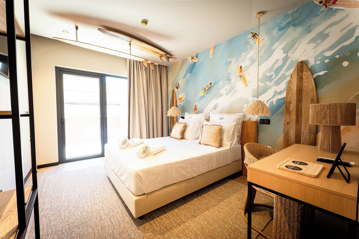 Una habitación de hotel con cama, escritorio, silla, un amplio ventanal y un mural en la pared.
