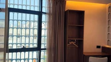 Habitación de hotel con cama, escritorio, silla, armario y un amplio ventanal con vistas a edificios.