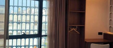 A hotel room with a bed, a desk, a chair, a wardrobe, and a large window with a view of buildings.