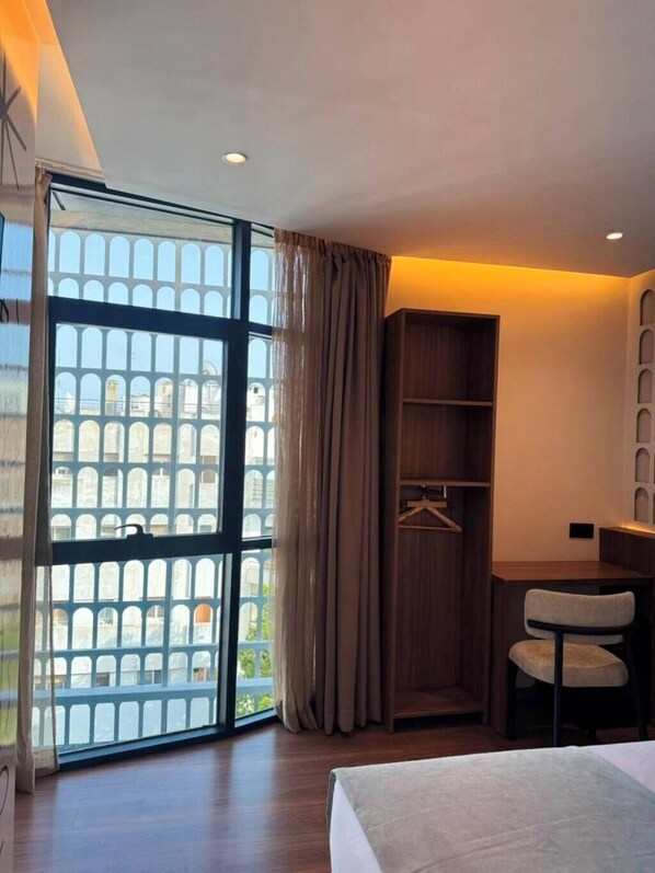 A hotel room with a bed, a desk, a chair, a wardrobe, and a large window with a view of buildings.