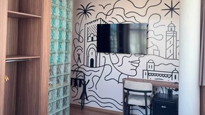 Habitación de hotel con armario de madera, separador de bloques de vidrio, televisor de pantalla plana montado en la pared, escritorio con silla y cama.
