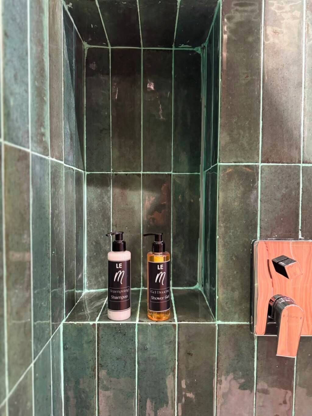 A bathroom with two bottles of Le M shampoo and shower gel, a wooden towel rack, and a green-tiled shower enclosure.