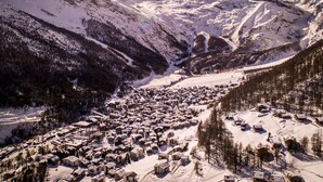 A snow-covered mountain village with buildings clustered along a valley, surrounded by forested slopes and mountains.