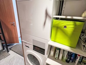 A compact, modern laundry room with a front-loading washing machine, a drying rack, and a shelf with various cleaning supplies.