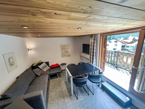 A modern living space with a sofa, a dining table with chairs, and a view of a residential area through large windows.