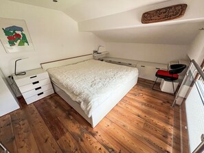 A bedroom with a bed, wooden flooring, a desk, and a chair.