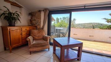 A room with a wooden cabinet, a cushioned chair, a small table, and a sliding glass door leading to an outdoor area with a view of hills.