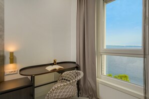 Een kamer met een gebogen houten tafel, een stoel, een boek, een wandlamp en uitzicht op zee door een raam met gordijnen.