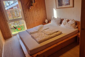A bedroom with a wooden bed, white bedding, a window with a view of trees, and a white brick wall. - Gemutlicher Doppelbungalow im Ferienpark Mirow mit Terrasse (Mirow)