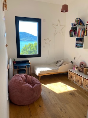 Et soveværelse med et stort vindue, en seng, et skrivebord, en poseleje og en reol.