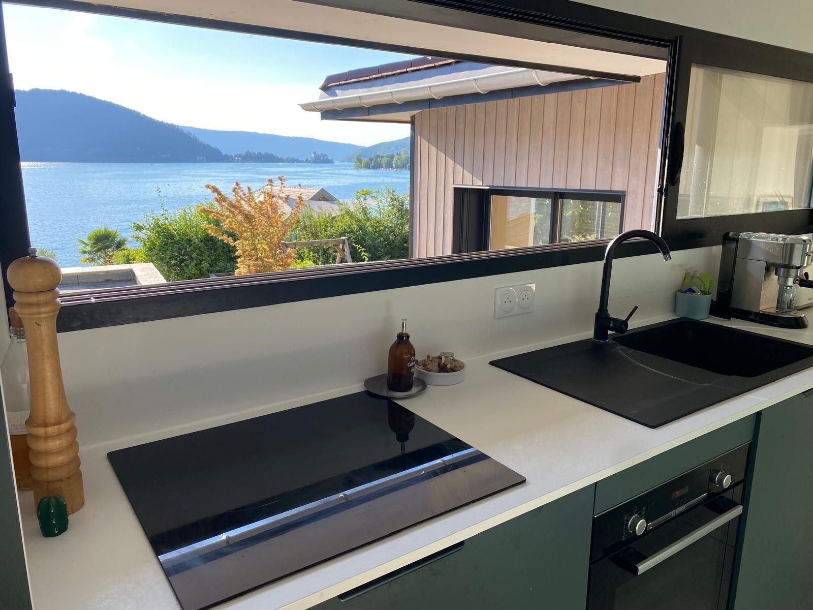 Eine moderne Küche mit großem Fenster, das auf einen See und Berge blickt, sowie eine Kaffeemaschine und ein Spülbecken.