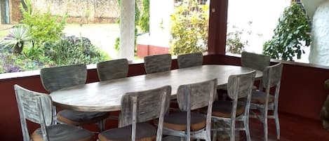 A covered outdoor dining area with a round table and chairs, surrounded by a red-tiled floor and white pillars.