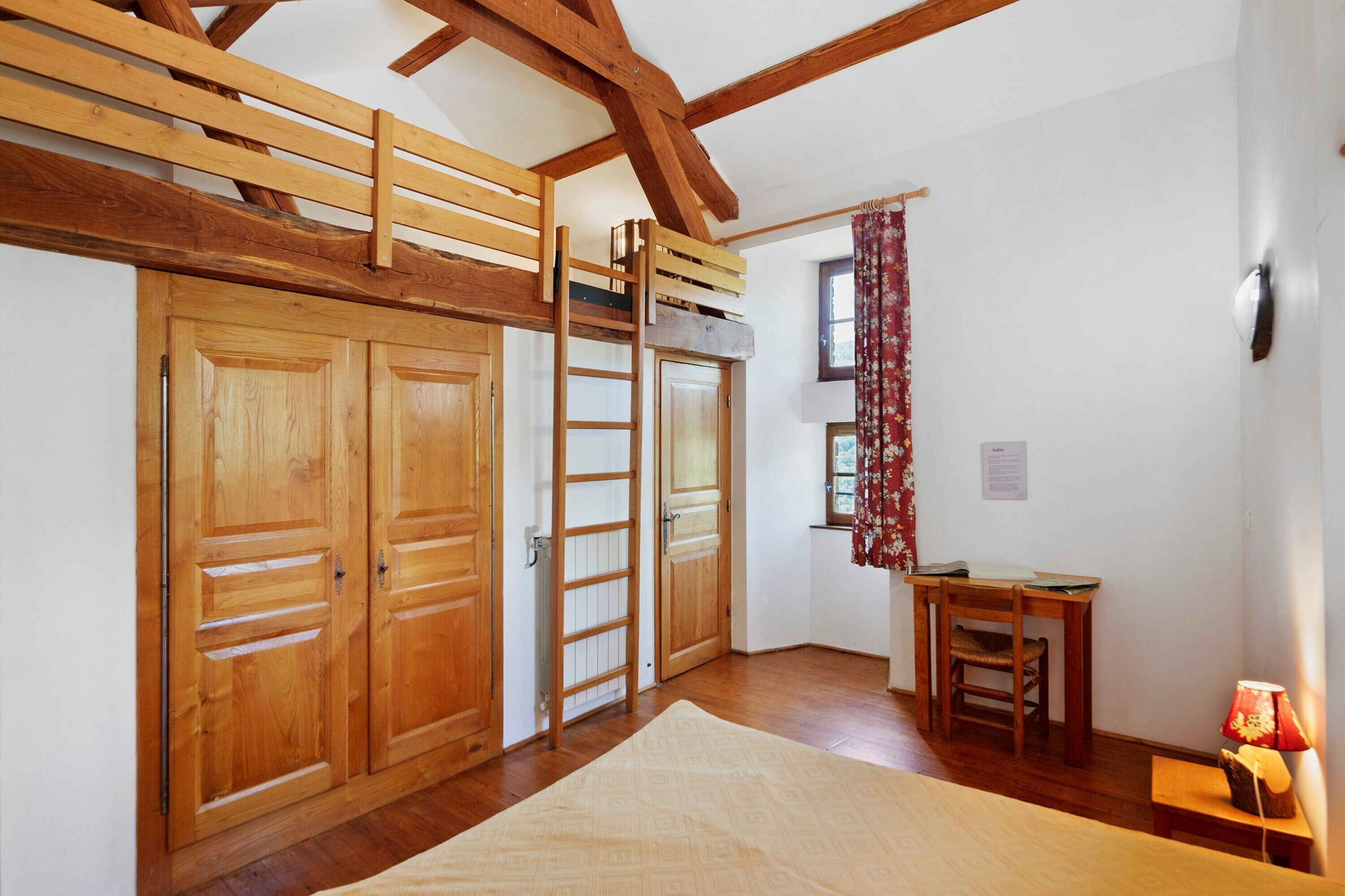 Een kamer met houten vloer, een hoogslaper, een bureau met stoel, een radiator en een raam met gordijnen.