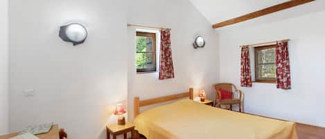 A room with a bed, a desk, two chairs, a window with curtains, and a ceiling with wooden beams.