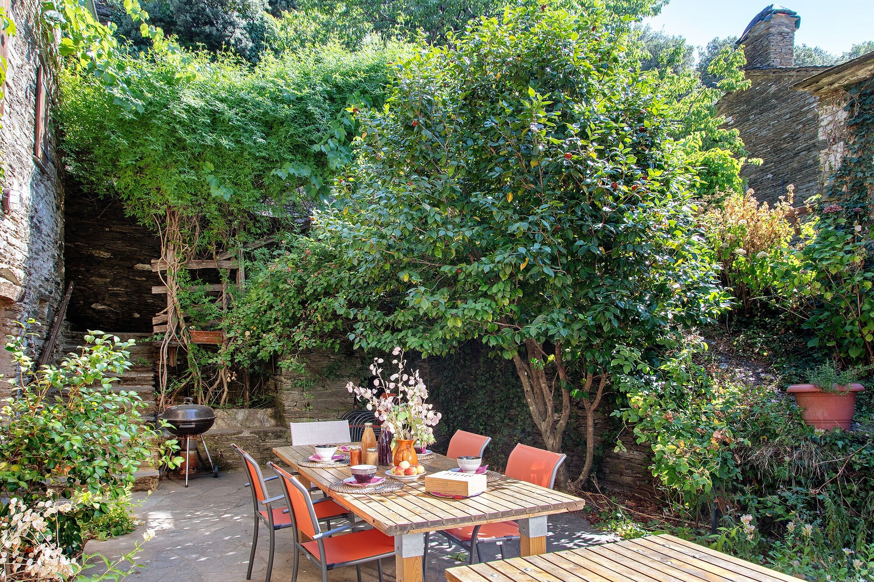 An outdoor wooden table set with chairs, surrounded by lush greenery and a stone wall.