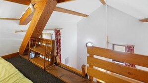 Ein Zimmer mit schräge Decke, Holzbalken, einem Etagenbett, einem kleinen Tisch und einem Fenster mit Vorhängen.