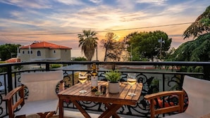 A wooden table with two wine glasses and a bottle, surrounded by white cushioned chairs, on a balcony with a view of the sea and buildings. - Studio Apartment 'Chrissi 103' with Sea View, Wi-Fi and Air Conditioning (Skala Sotiros)