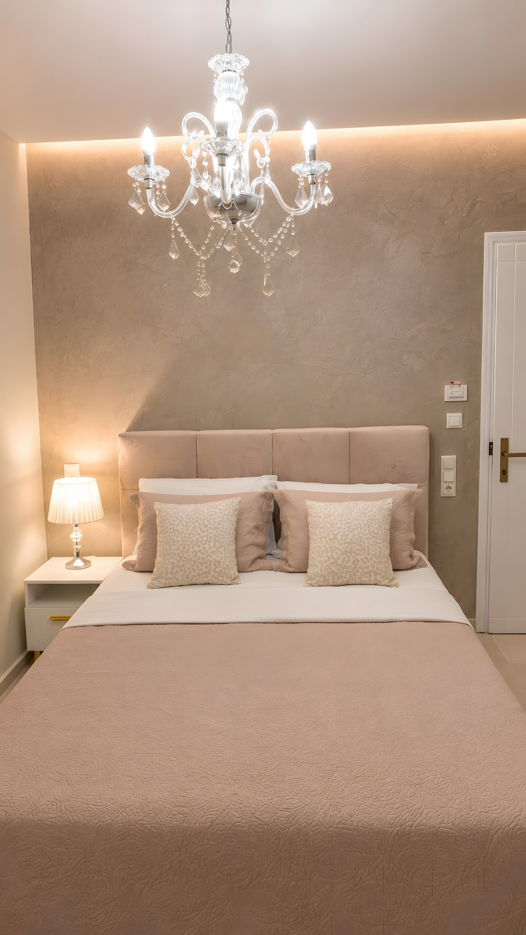 Een slaapkamer met een groot bed, een nachtkastje met lamp, een kroonluchter en een deur.