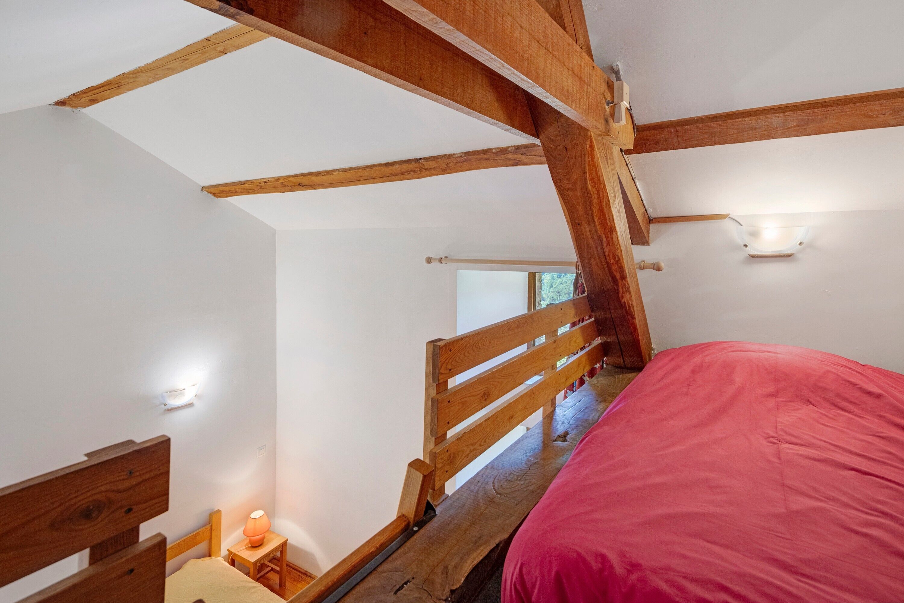 A bedroom with a wooden bed, a nightstand with a lamp, and a window with a view of trees.
