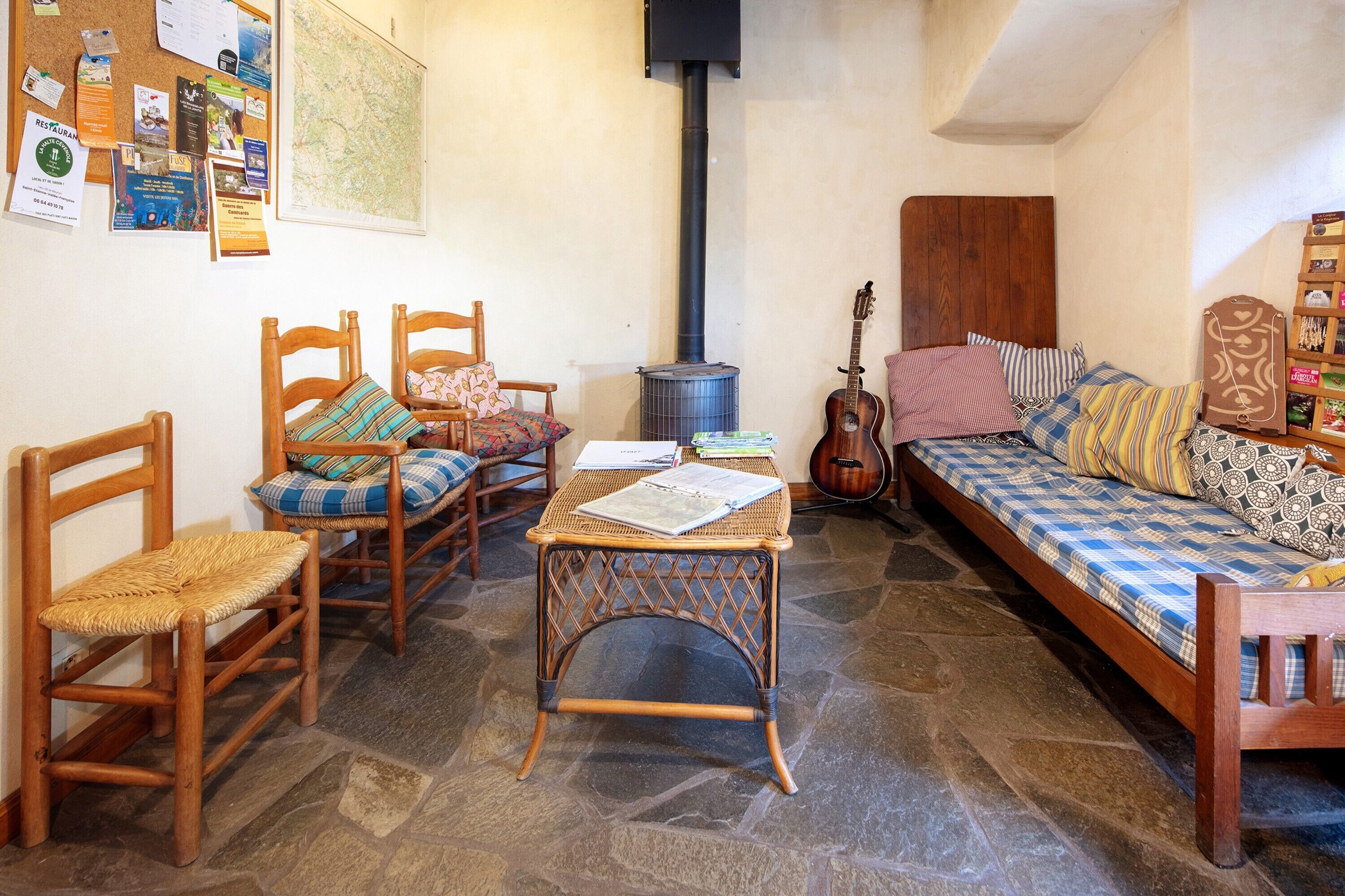 Une pièce chaleureuse avec un sol en pierre, un lit en bois recouvert d’une literie à motifs, une guitare, une table avec des livres et un mur où divers objets sont accrochés.