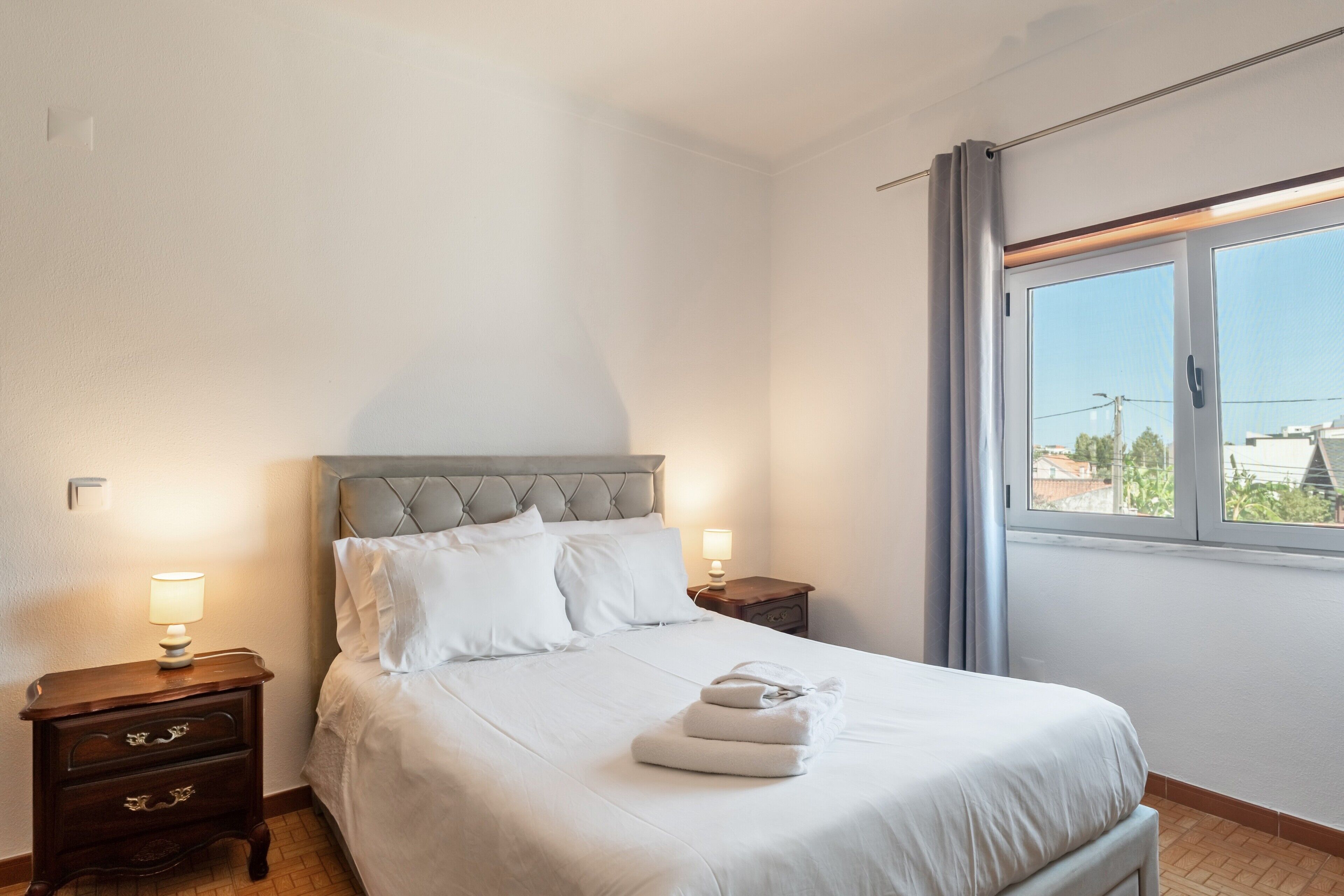 Una cama bien hecha con ropa blanca, una mesita de noche de madera con una lámpara, y una ventana con vistas a edificios.