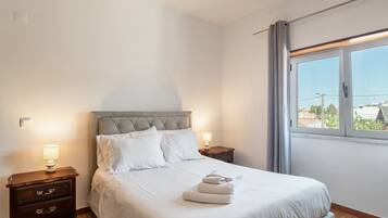 Una cama bien hecha con ropa blanca, una mesita de noche de madera con una lámpara, y una ventana con vistas a edificios.