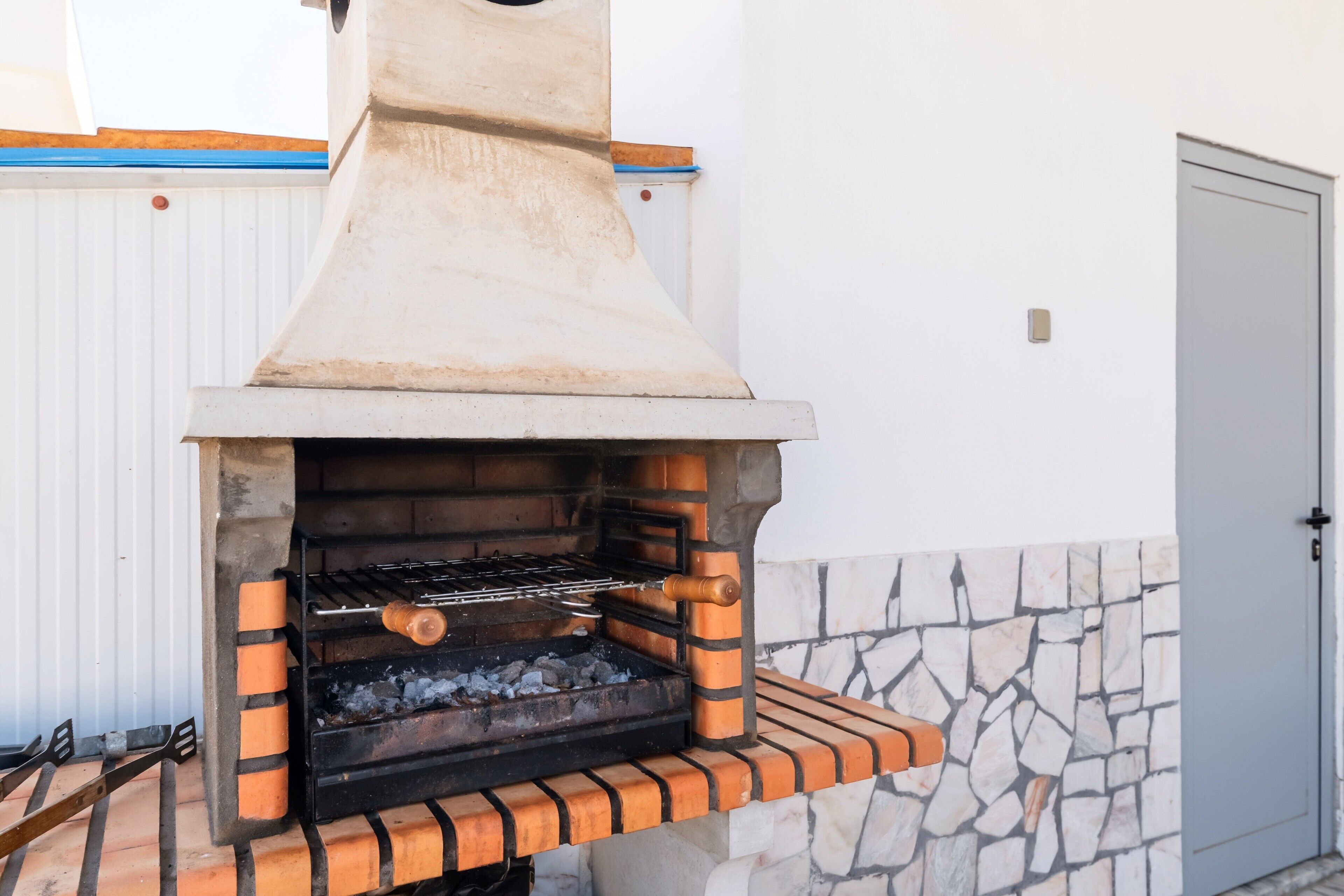 Un barbecue extérieur en briques, doté d’une cheminée, est installé contre un mur blanc près d’une porte.