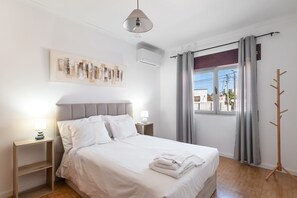 A neatly made bed with white linens, a bedside lamp, and a wall-mounted air conditioning unit. - Holiday Home 'Vivenda Alves' with Mountain View, Wi-Fi and Air Conditioning (Charneca da Caparica)