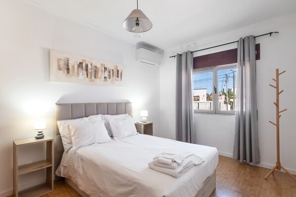 A neatly made bed with white linens, a bedside lamp, and a wall-mounted air conditioning unit.