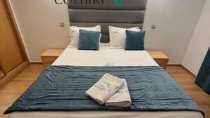 Ein ordentlich bezogenes Bett mit blauer Bettdecke und Kissen, ein gefaltetes Handtuch mit Logo und ein Logo über dem Bett.