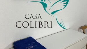 Ein Wandbild zeigt einen Strandweg, ein Logo mit einem Vogel und ein blaues Handtuch mit der Aufschrift 'Casa Colibri'.