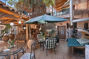 Een terras buiten met een tafel en stoelen, een grote parasol en diverse potplanten.