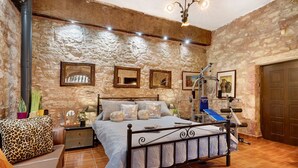 Un dormitorio con pared de piedra, una cama con cabecera, una mesita de noche, una lámpara y una puerta.
