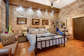 Un dormitorio con pared de piedra, una cama con cabecera, una mesita de noche, una lámpara y una puerta.