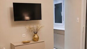 A bedroom with a bed, a TV mounted on the wall, a small table with a vase, and a chandelier. - Studio Apartment 'Chrissi 105' with Mountain View, Wi-Fi and Air Conditioning (Skala Sotiros)
