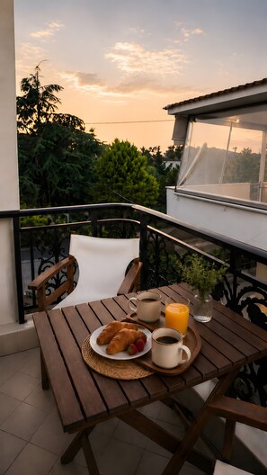 A wooden table with breakfast items, a chair, and a view of trees and a sunset. - Studio Apartment 'Chrissi 105' with Mountain View, Wi-Fi and Air Conditioning (Skala Sotiros)