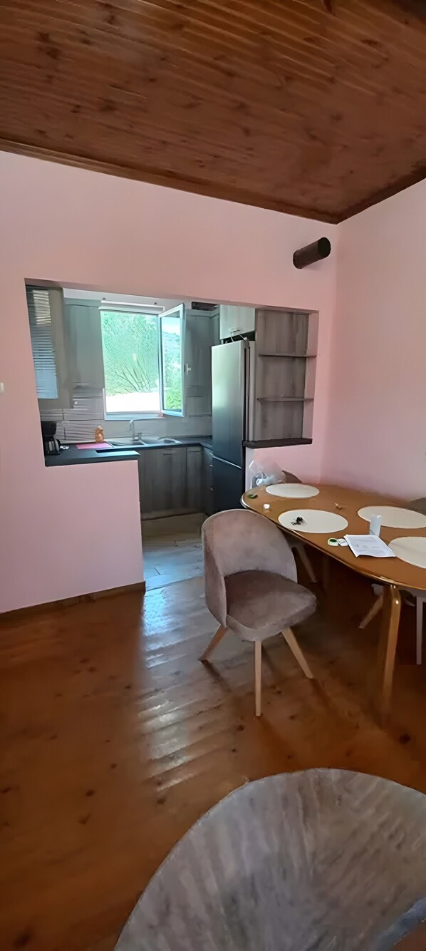 A room with a wooden floor, a dining table with chairs, a kitchen area with a sink, and a window with a view of greenery.
