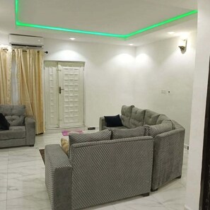 Una sala de estar con un sofá modular, un sofá más pequeño y una mesa de centro. Hay una puerta con un panel de vidrio y una cortina.
