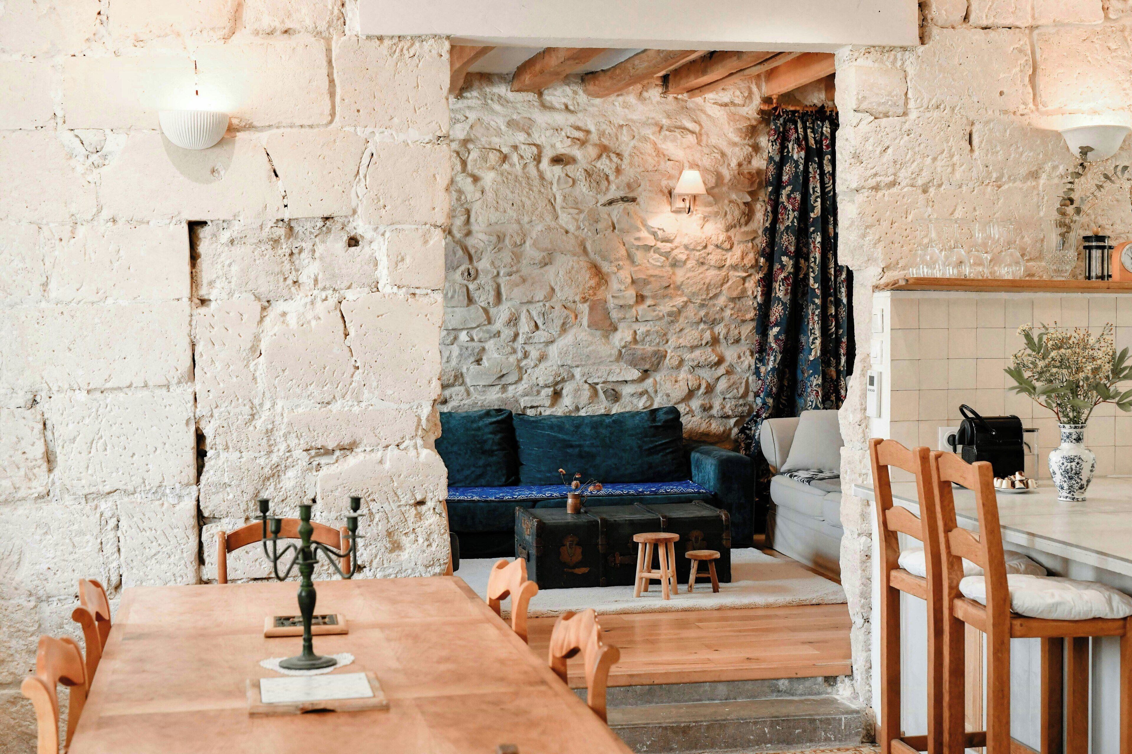 Un intérieur rustique avec des murs en pierre apparentes, une table à manger en bois, un canapé et un coin cuisine agrémenté d’un pare-feu en carreaux.