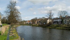 A river with a wooden walkway and stone wall on one side, surrounded by traditional buildings with multiple stories and pitched roofs. - Riverside Escape - Newark on Trent (Newark)