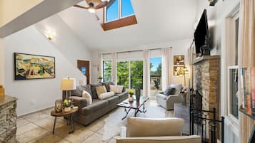 A spacious living room with a stone fireplace, a large window with a view of greenery, and a ceiling fan.